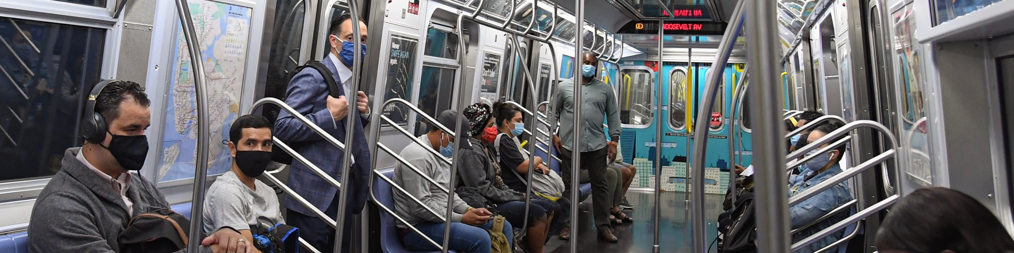 People wearing masks sit and stand on a subway train. The car isn’t crowded and there are a few empty seats.