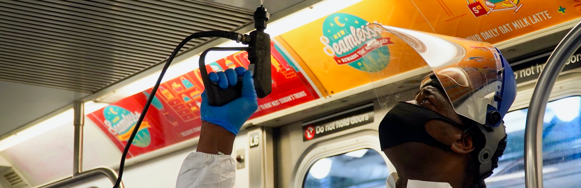 A man with a mask and face shield sprays cleaning solution onto the ceiling of a subway train.