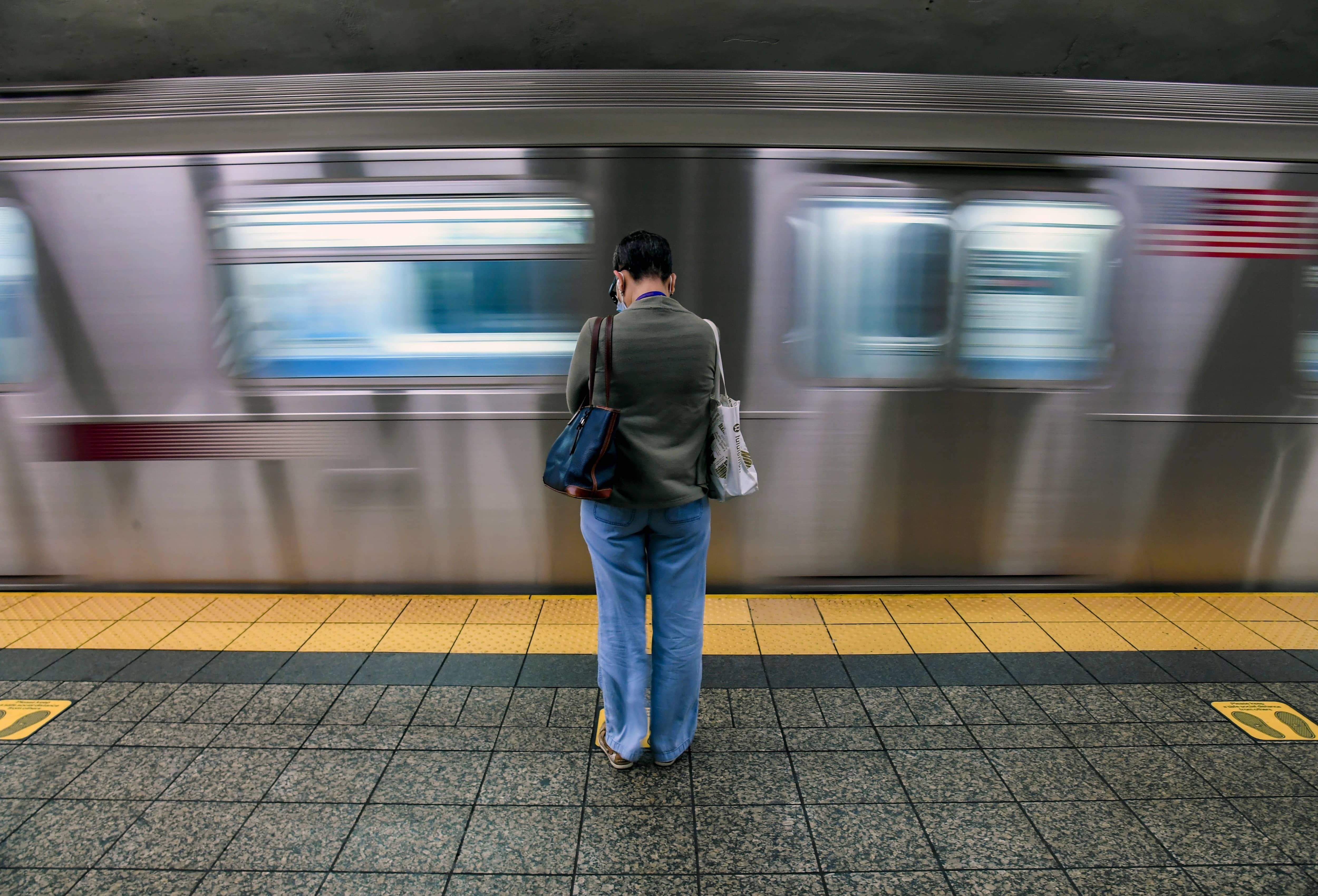 A person in a sage-green jacket and jeans stands on a subway station platform. An arriving train is visible in the background, blurred because of the motion.