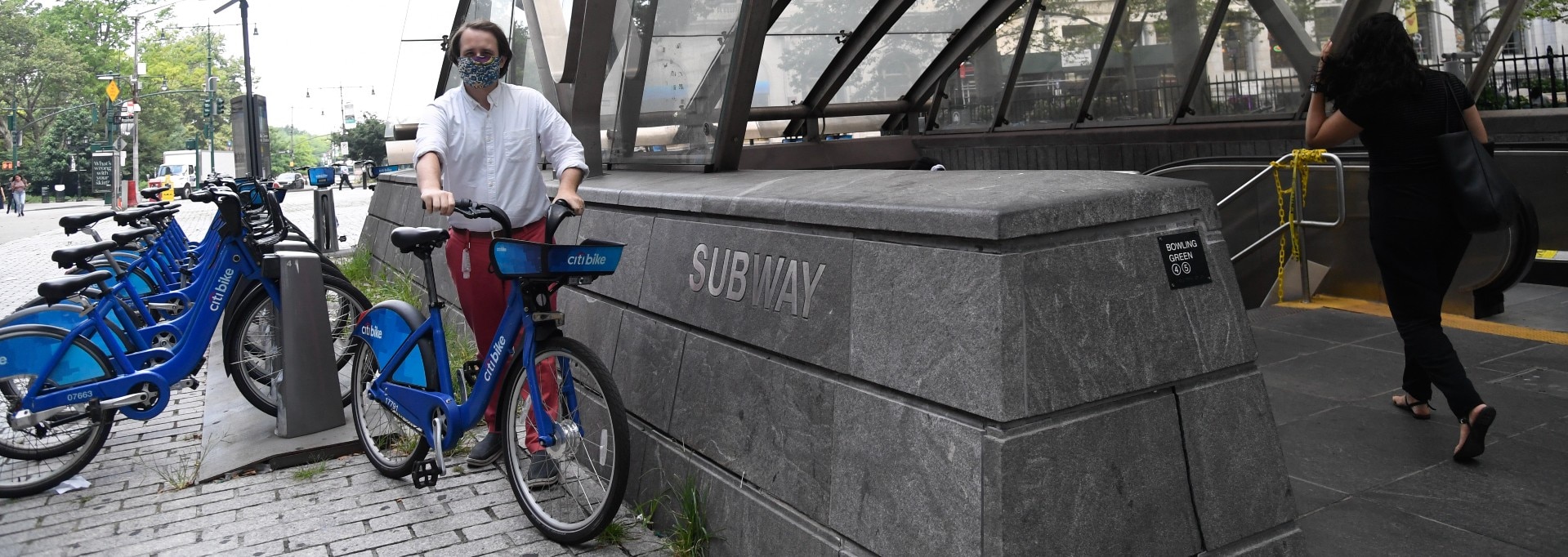 A large blue bike being wheeled by a man wearing red pants. He is next to a subway station entrance. 