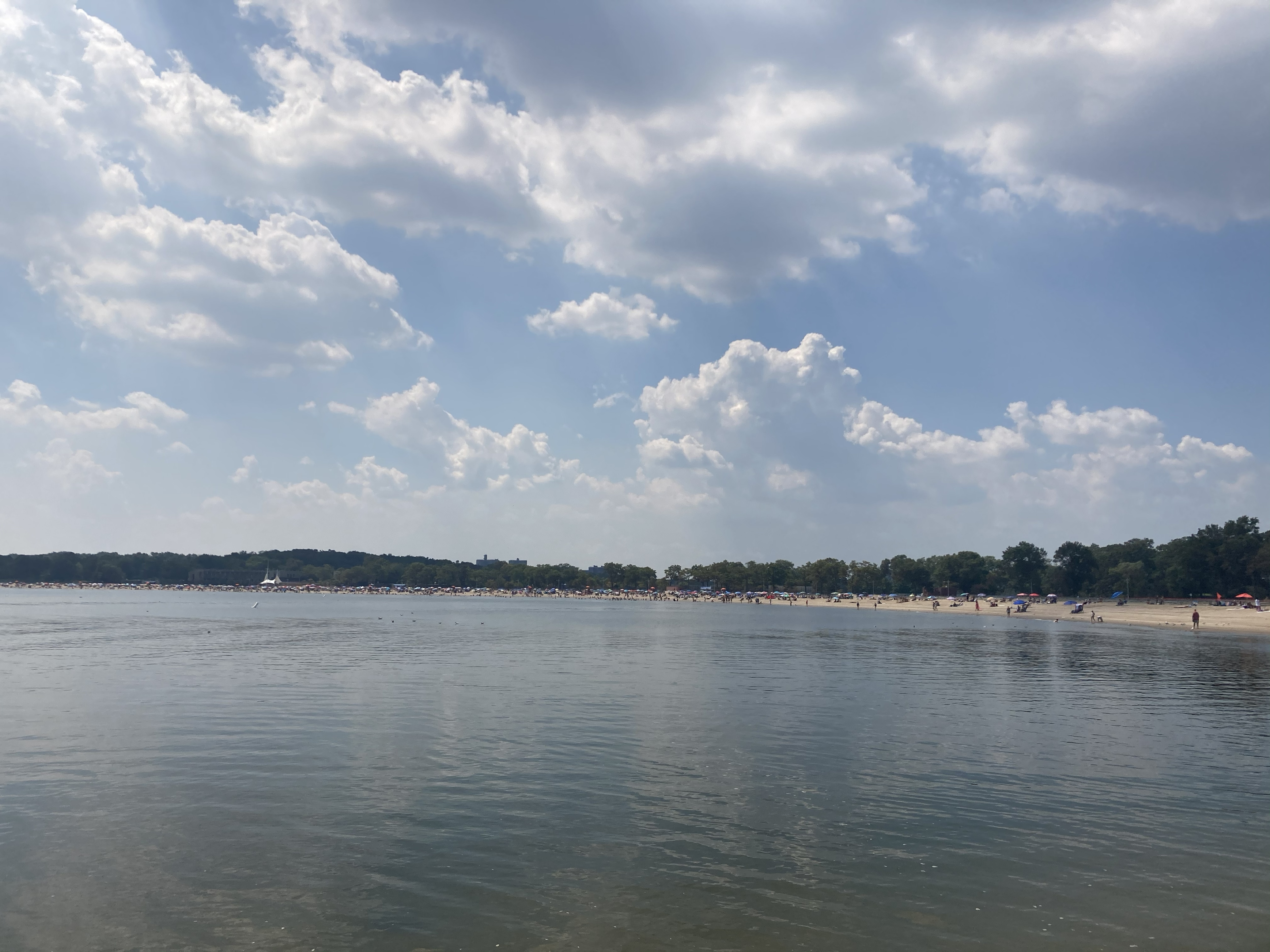 Wide shot of calm water at Orchard Beach, under a bright blue sky with puffy white clouds, and a moderate amount of people on the beach at the right.