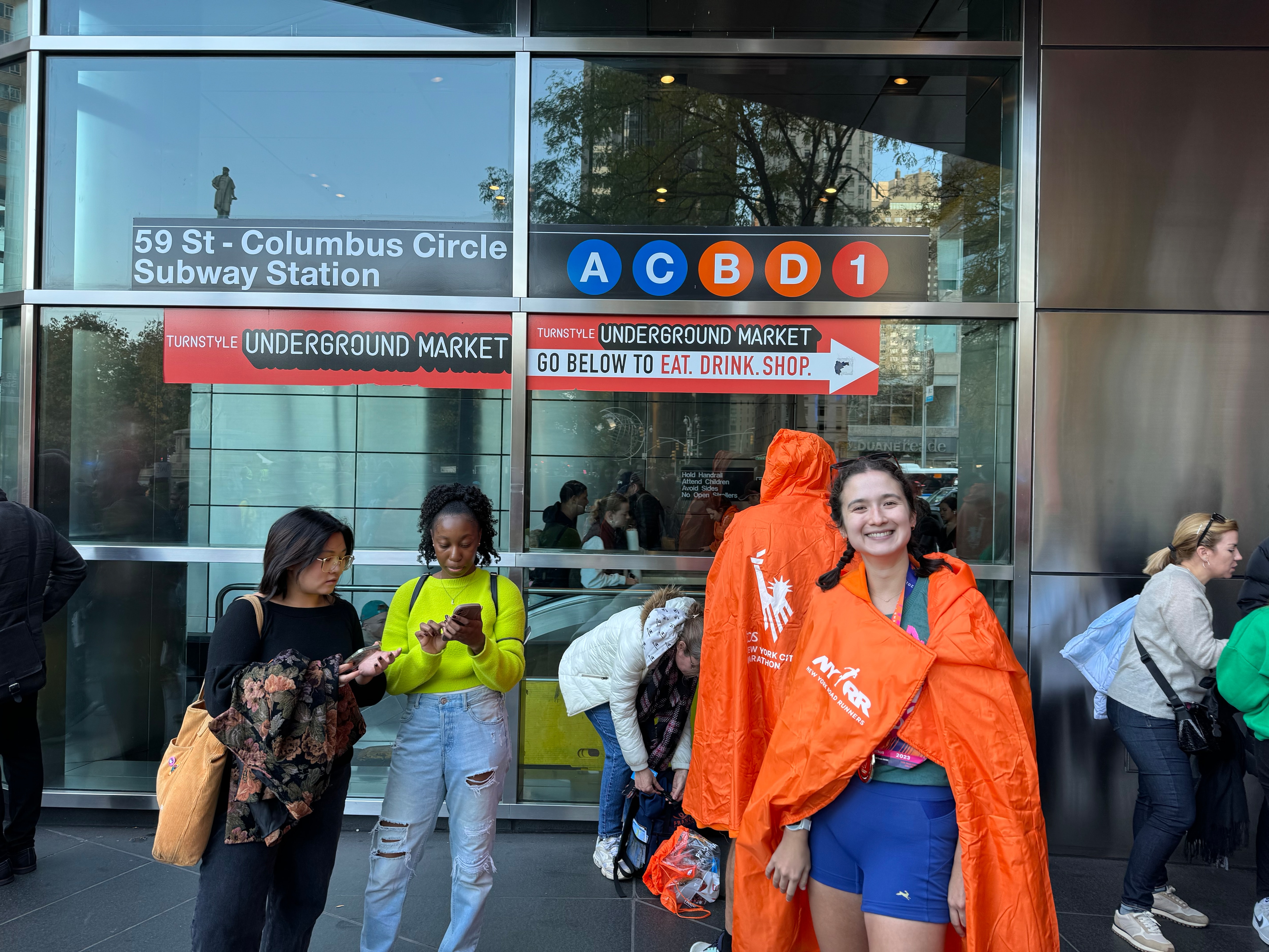 Lisa Mae Fiedler smiling and wearing an orange New York Road Runners poncho in front of the 59 St-Columbus Circle A C B D 1 subway station after finishing the 2023 New York City Marathon.