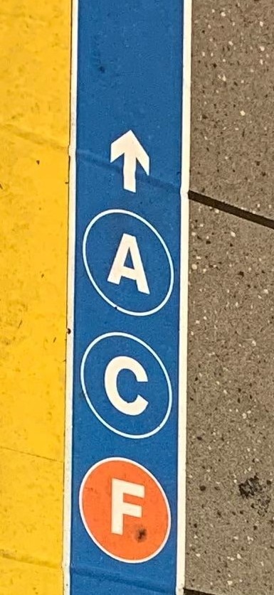 blue tape on station floor has small white border and the A, C, and F train bullets and arrow pointing forward.