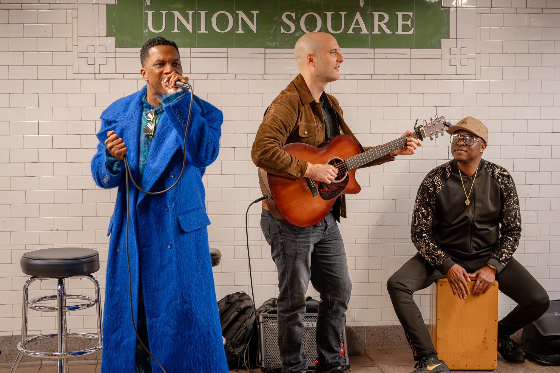 Leslie Odom Jr., wearing a bright blue coat, sings as a guitarist and drummer play behind him at the Union Square subway station, with the Union Square tile sign behind them