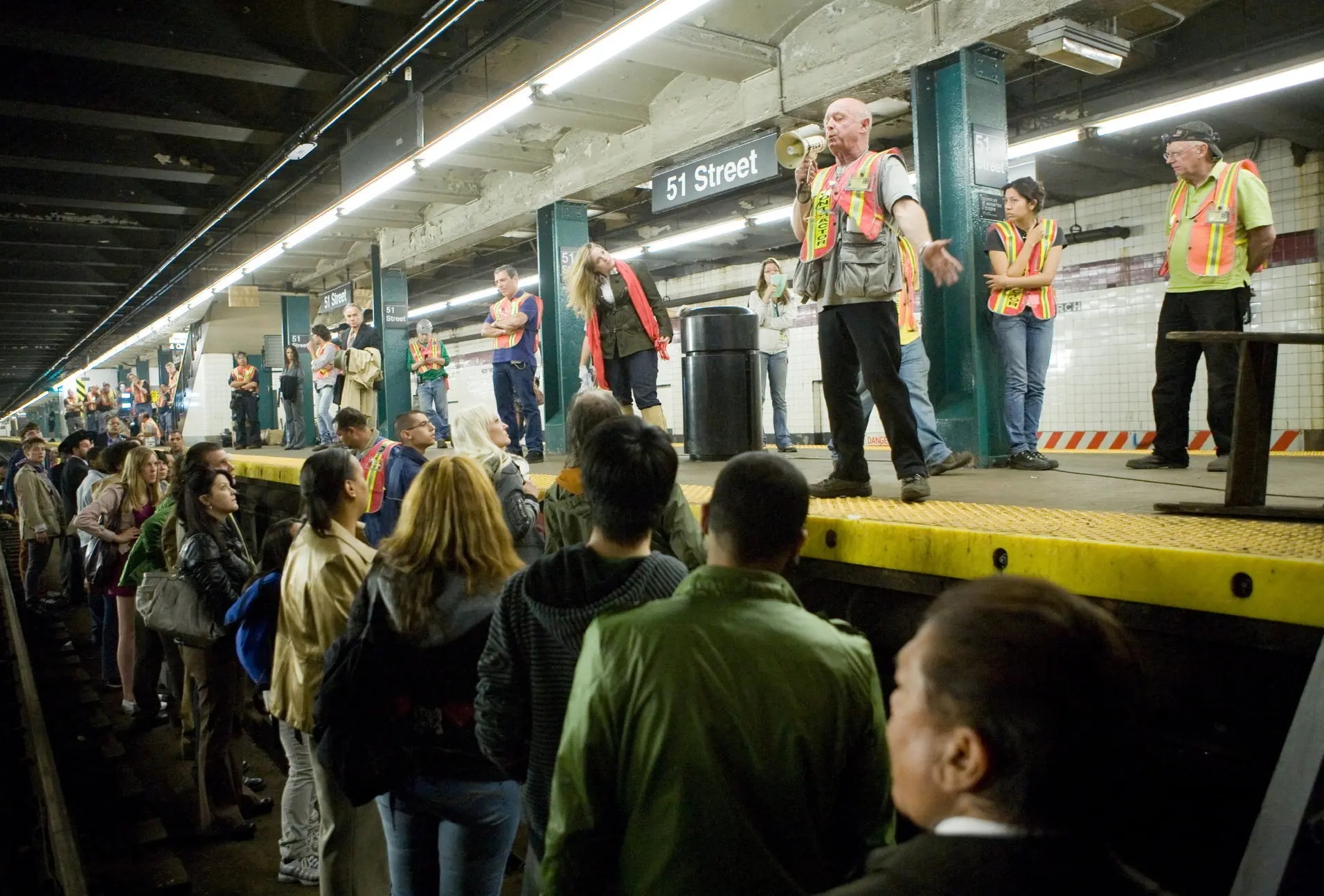 Film production members wearing safety vests are standing on a platform, one with a bullhorn, speaking to actors and extras who are standing in the train tracks