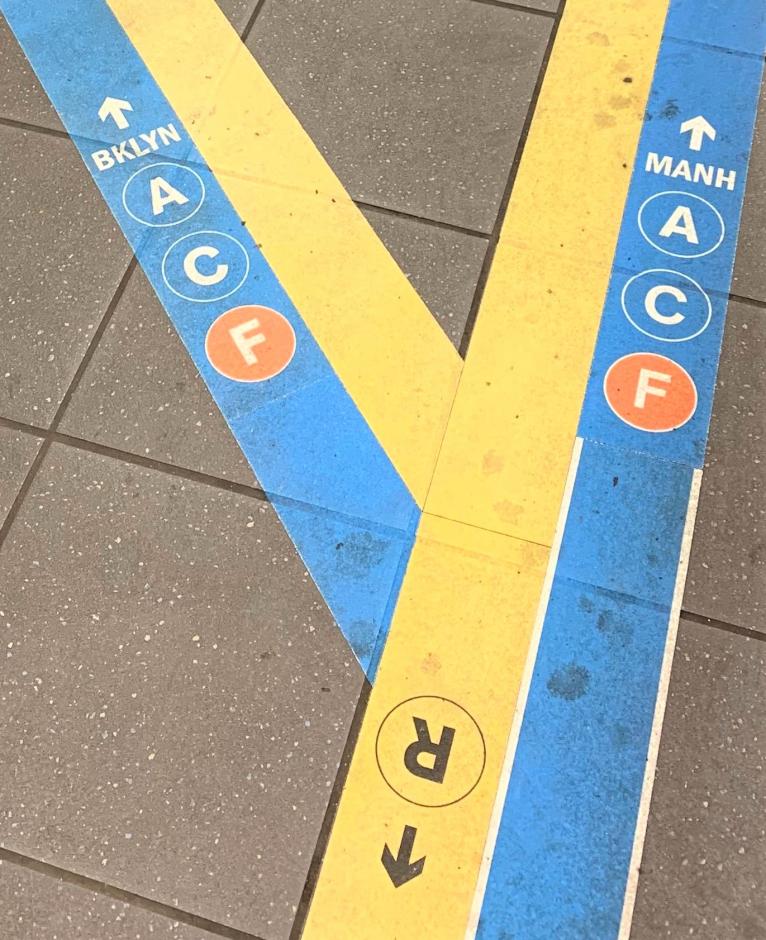 brightly colored tape on station floor with yellow tape with upside-down R train bullet pointing behind, and a Y-shaped split from the R tape where two pieces of blue tape, each with the A, C, and F train bullets divert in saying "BKLN" and "MANH" and arrows pointing diagonally left and diagonally right.