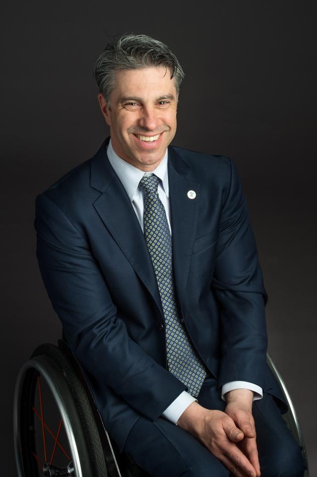 A photo of a man in a suit and tie, smiling and sitting in a wheelchair with his hands in his lap.