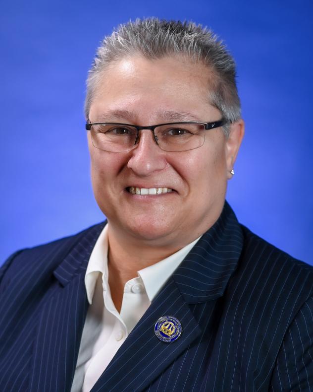A woman with short gray hair wears a white button-down shirt and a navy blue blazer. She is wearing glasses and is smiling.