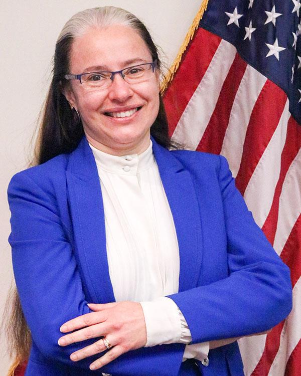 A headshot of Lisa Schreibman wearing a blue jacket and a white shirt with an American flag in the background