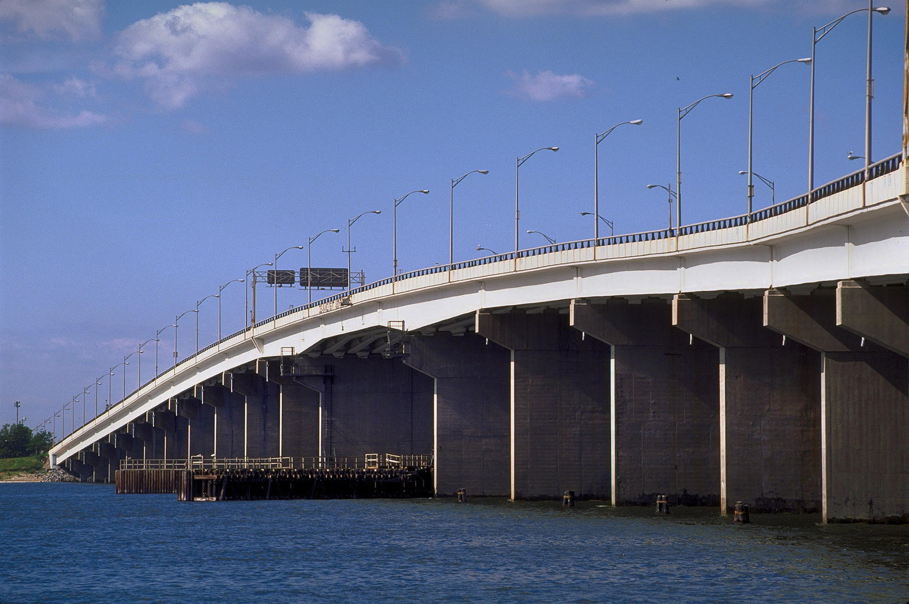 A bridge with traffic signs and streetlights visible stretches across a body of water, with blue sky and clouds overhead.