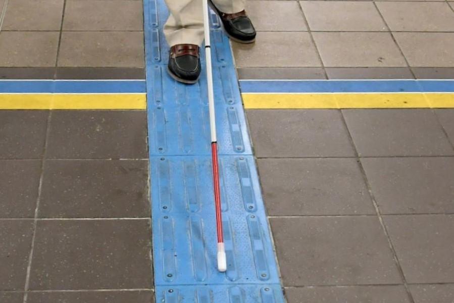 A person with a red-tipped white cane navigates a subway station using blue raised floor tiles.