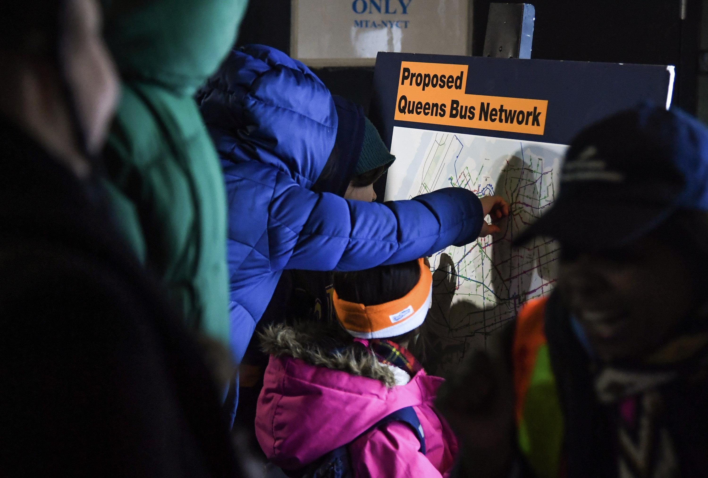 A small crowd of people gather around a large poster that reads, "Proposed Queens Bus Network." A small child is reaching out to touch the poster.