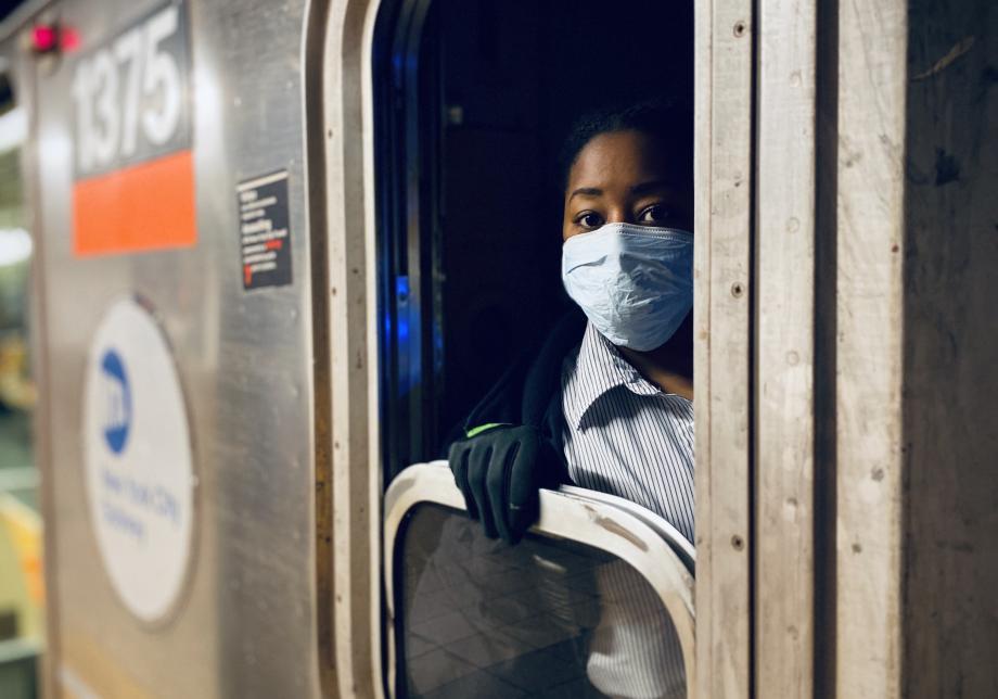 A female train operator wearing a face mask and gloves looks out from inside a subway train.