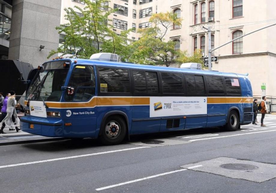 A large bus parked along a street in Manhattan. The bus is branded with blue and gold MTA colors. 