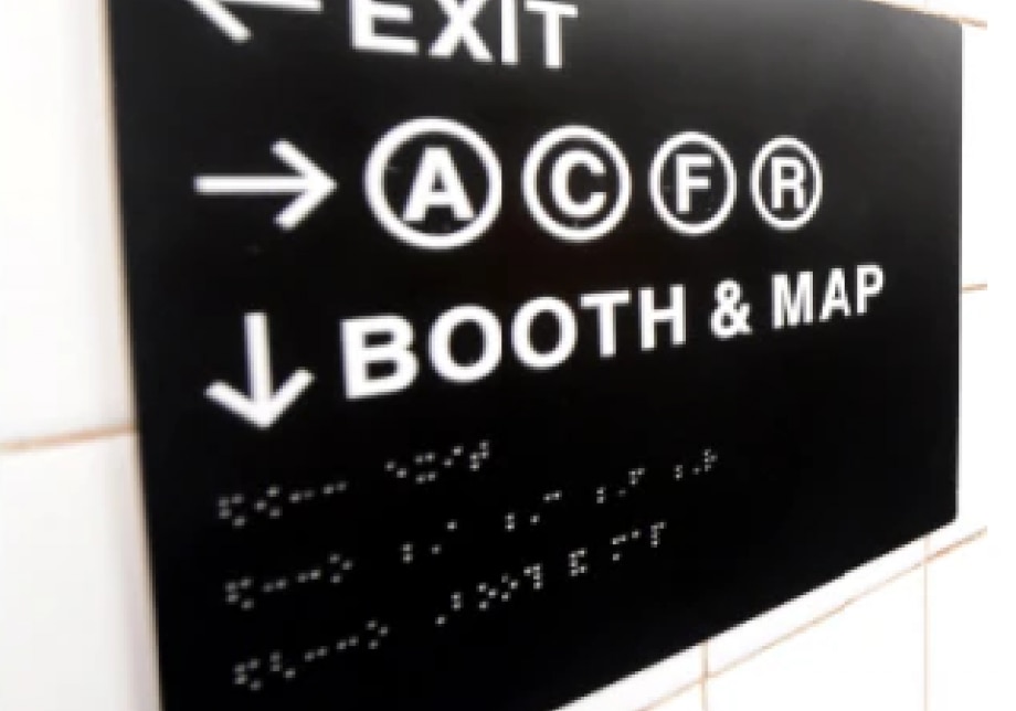 A Braille/raised lettering sign reading: [left arrow] "exit," [right arrow] A C F R, [down/forward arrow] "booth & map."