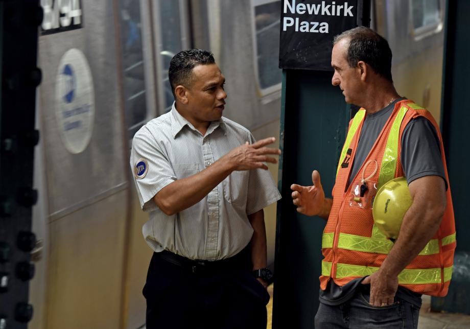 Two MTA employees, one in a short-sleeved collared shirt and the other in an orange construction vest and a hard hat, talk on a subway platform. A train is visible behind them.
