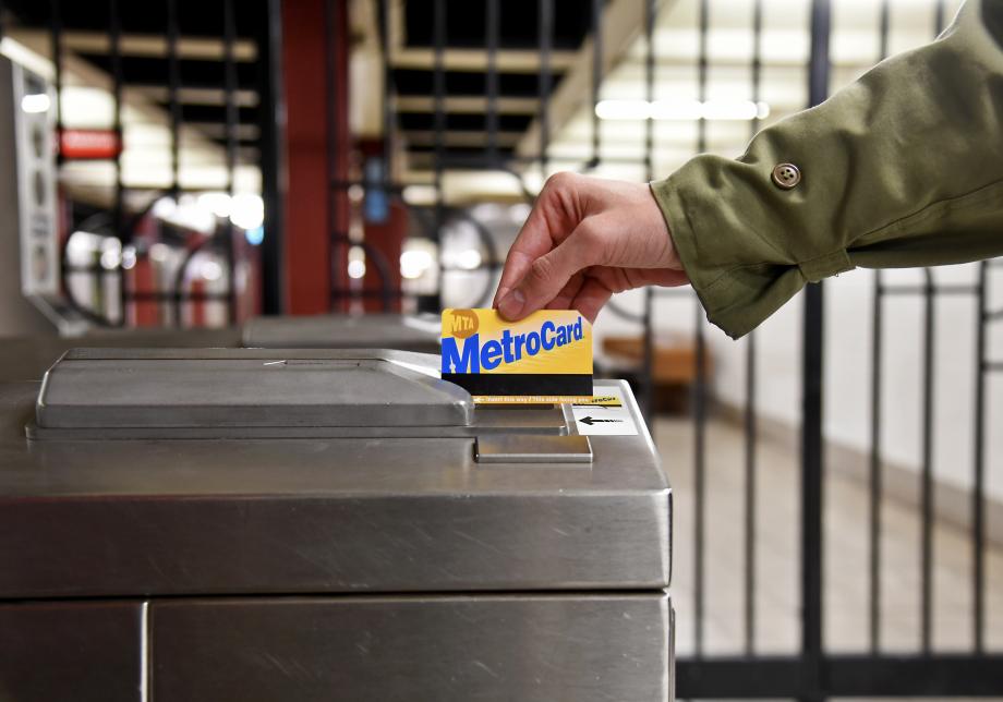 A closeup of someone swiping a MetroCard at a turnstile. The blue and gold MetroCard logo is facing toward the camera and the person swiping it, with the black barcode visible at the bottom. 