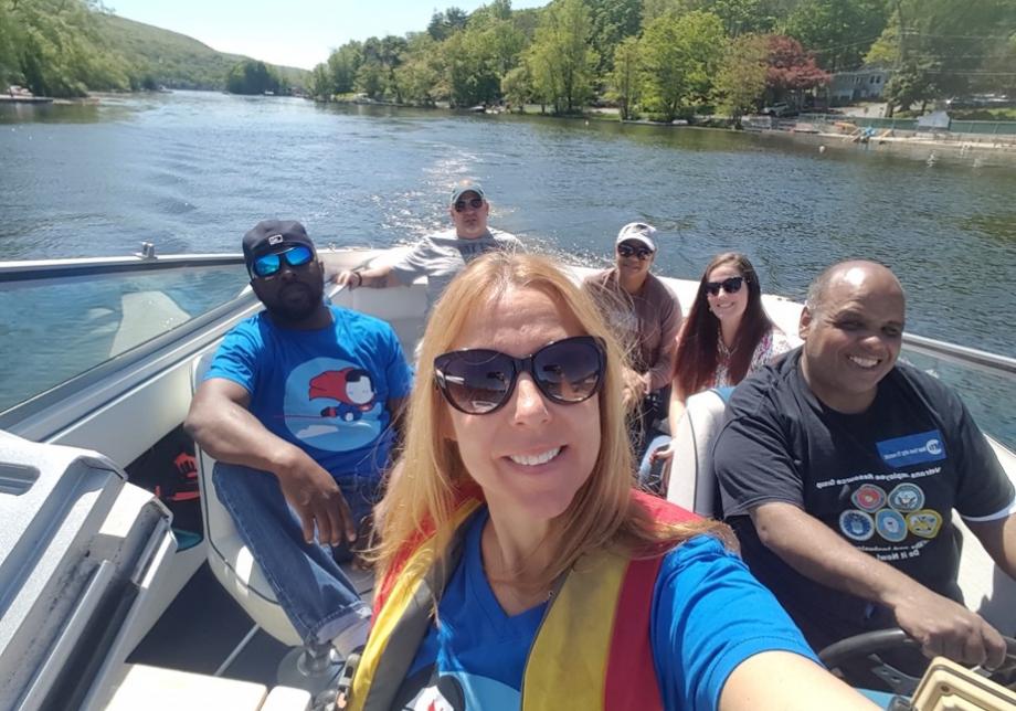 A woman in sunglasses takes a photo of herself and five other people in a boat on the water. Everyone is smiling, and one man is wearing a T-shirt that says Veterans Transit Employee Group.