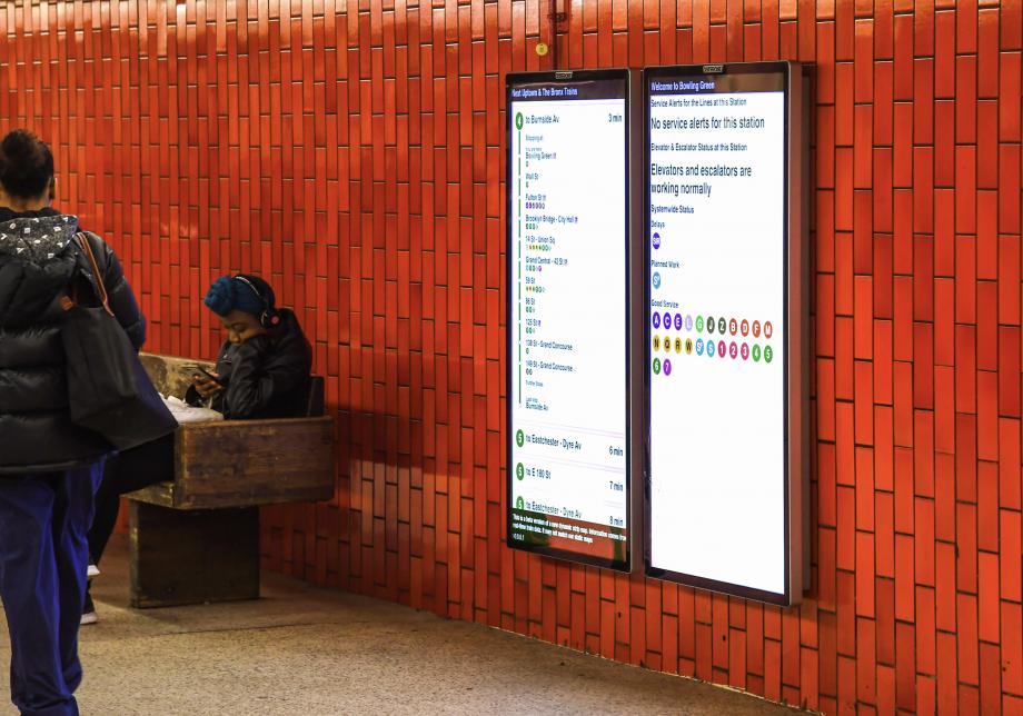 People sit near and walk by two large digital screens on the wall of a subway station. The screens show arriving trains and service status.