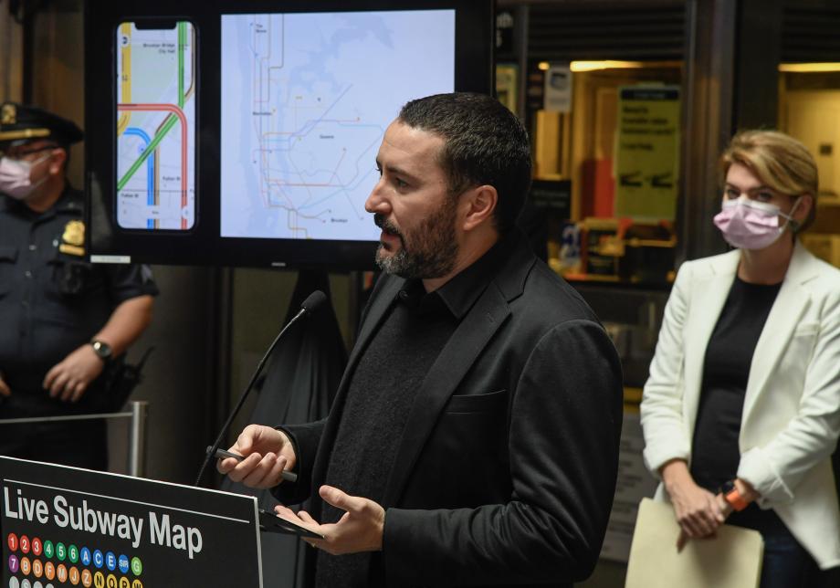 A man stands at a lecturn in a subway station. A screen with a subway map is visible over his shoulder.
