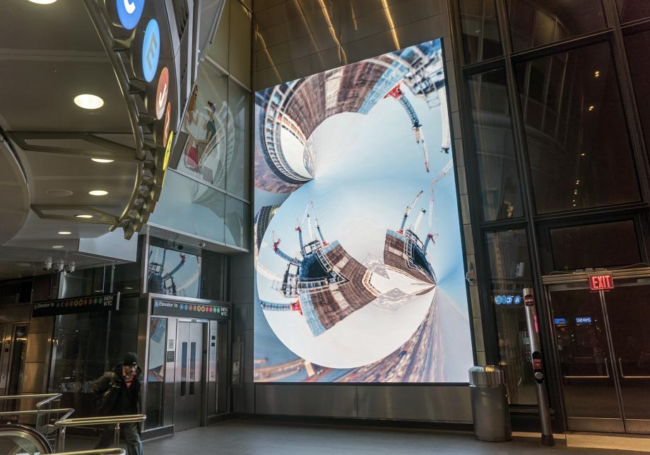 Anne Spalter_01  	  The photo shows digital artwork, New York Dreaming, created by Anne Spalter at Fulton Center. Large vertical screen displays a distorted view of a city scape.     