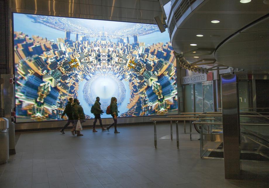 The photo shows digital artwork, New York Dreaming, created by Anne Spalter at Fulton Center. Large horizontal screen displays a distorted view of a city scape. 