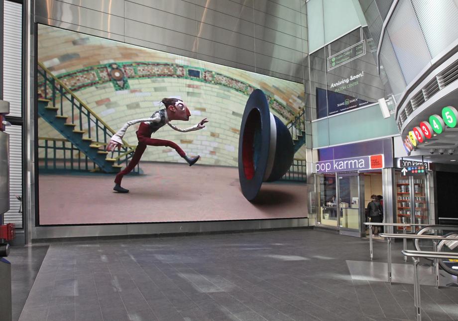 The photo shows digital artwork, The Bowling Bowler, created by Chris Sickels at Fulton Center. Large horizontal screen displays a sculpted figurine stepping into a large hat. 