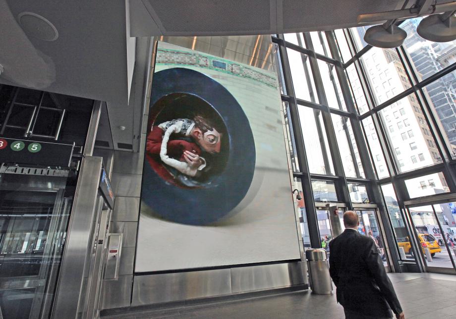 The photo shows digital artwork, The Bowling Bowler, created by Chris Sickels at Fulton Center. Large vertical screen displays a sculpted figurine traveling in a large hat. 