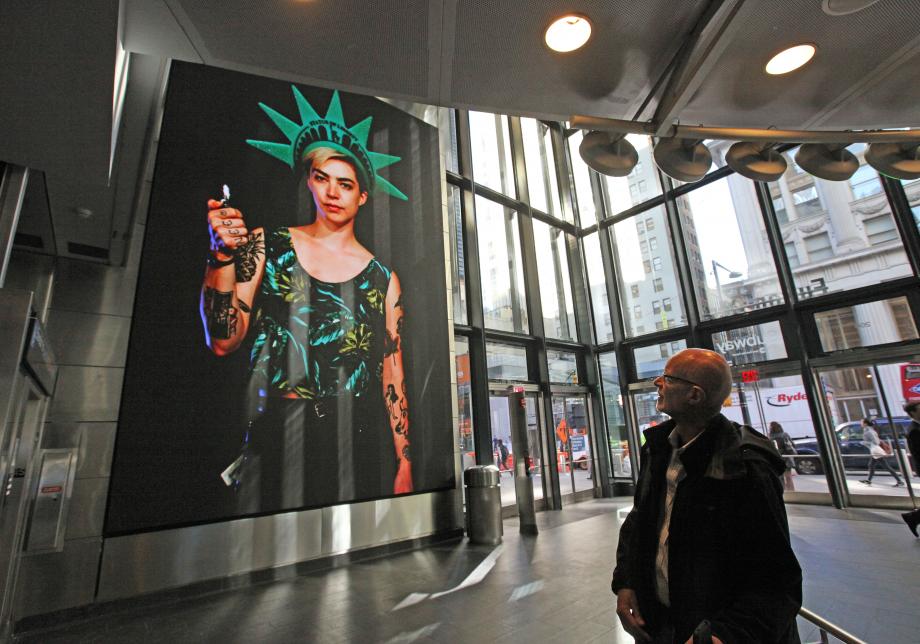 The photo shows digital artwork, New York Minute, created by Gabriel Barcia-Colombo at Fulton Center. Large vertical screen displays a woman with a  Statue of Liberty crown on a black background.  