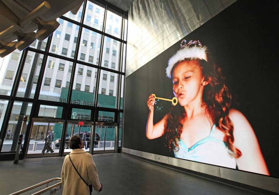 The photo shows digital artwork, New York Minute, created by Gabriel Barcia-Colombo at Fulton Center. Large horizontal screen displays a child blowing bubbles on a black background.  