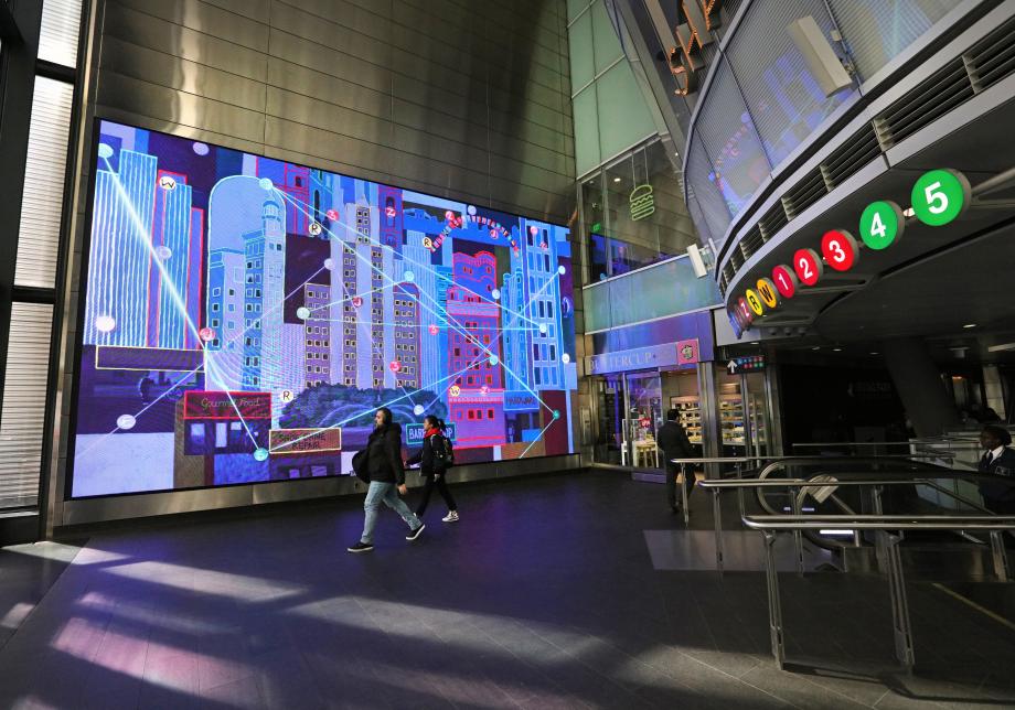 The photo shows digital artwork, Fulton Flow, created by Ezra Wube at Fulton Center. Large screen displays layered buildings, lines and colors.  
