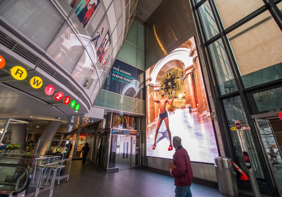 The photo shows digital artwork, Set Pieces, created by Rashaad Newsome at Fulton Center. Large vertical screen displays a man with red shoes in an imagined space.  