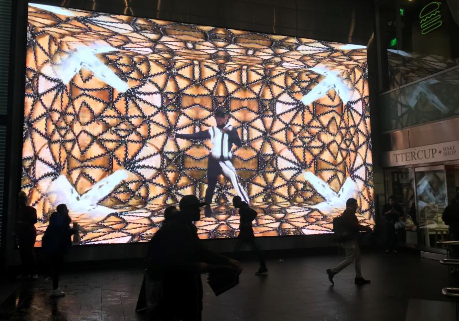 The photo shows digital artwork, Set Pieces, created by Rashaad Newsome at Fulton Center. Large horizontal screen displays a man with black and white clothing in a patterned space.  