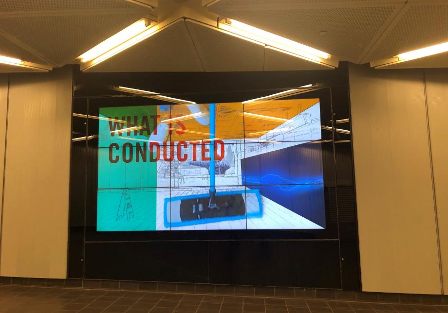 The photo shows digital artwork, WHAT IS HAPPENING, created by Sarah Rothberg & Marina Zurkow at Fulton Center. Large horizontal screen displays a man with black and white clothing in a patterned space.  