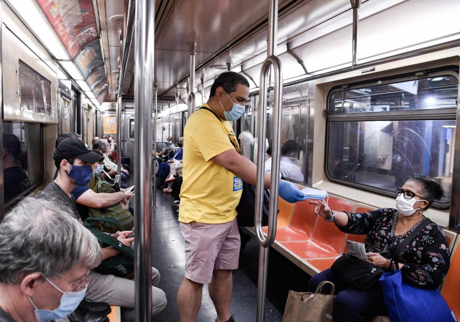 A man wearing a face mask hands a mask to a woman on a subway car. 