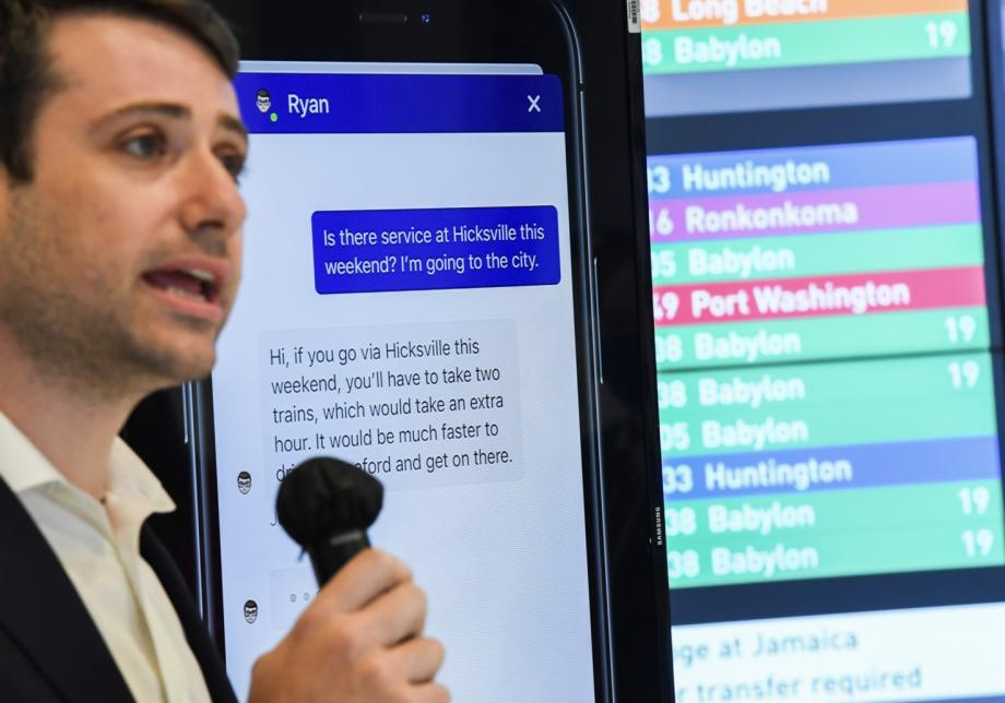 A man with a microphone speaks in front of digital screens that are showing information from an app. 