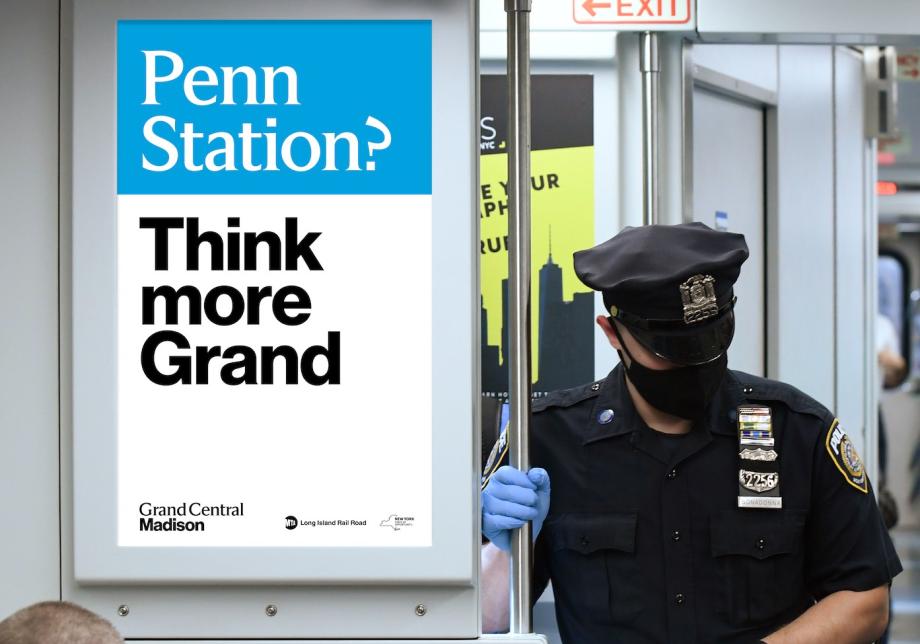 A police officer in uniform stands next to a sign on a train. The sign reads "Penn Station? Think more grand"