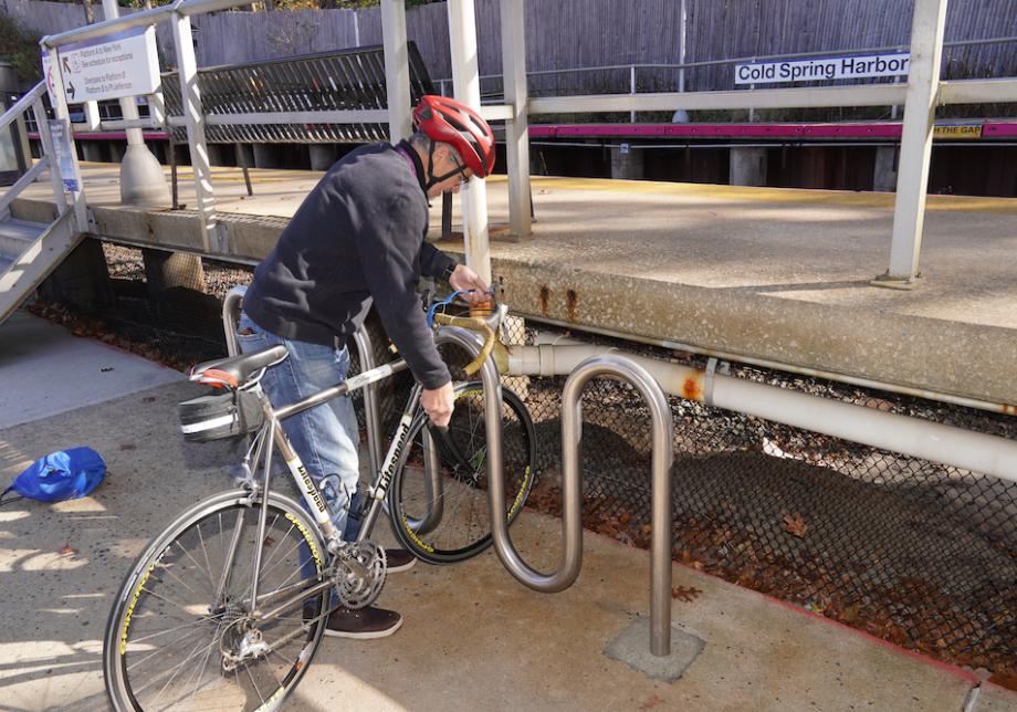A man in a red helmet locks a bicycle to a bike rack outside of a train station.