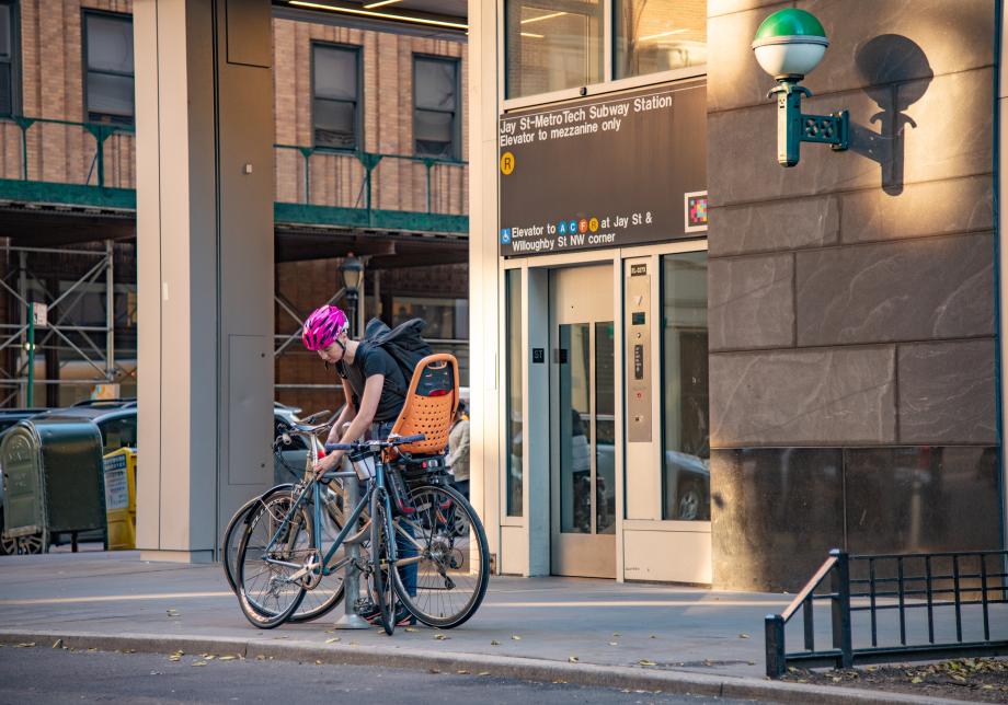 A man in a pink helmet locks his bike to a rack outside of a subway station. 