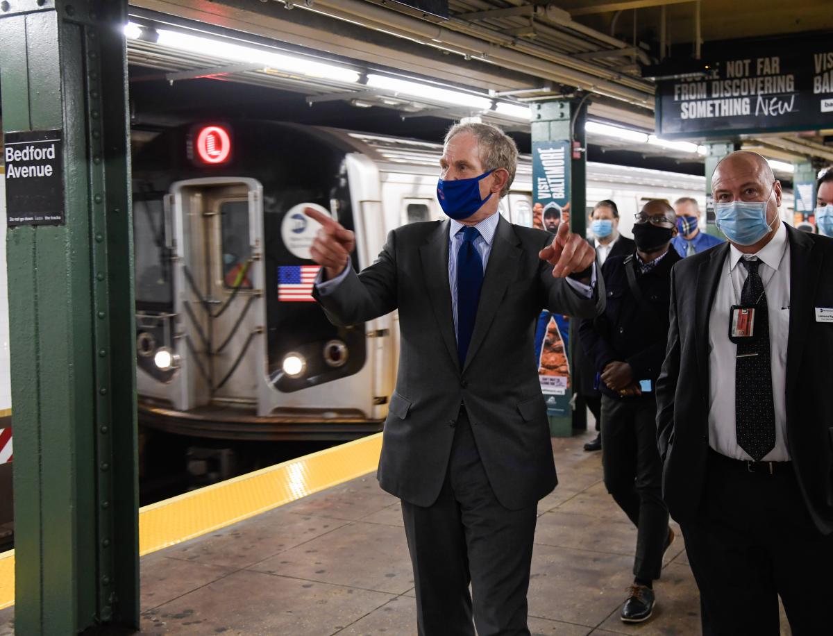 Janno Lieber, wearing a suit and tie and a face mask that says "New York Tough" on it, walks along a subway platform. He is gesturing to either side of the platform while an L train pulls into the station on one side. Other officials walk behind him. 