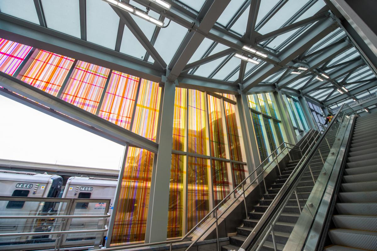 A permanent glass art installation by artist James Little on escalator wall interior at LIRR Jamaica Station shows colorful intricate lines that together form flat gradated blocks of color in the colors of the rainbow, from the perspective of the bottom of the escalator and stairs.