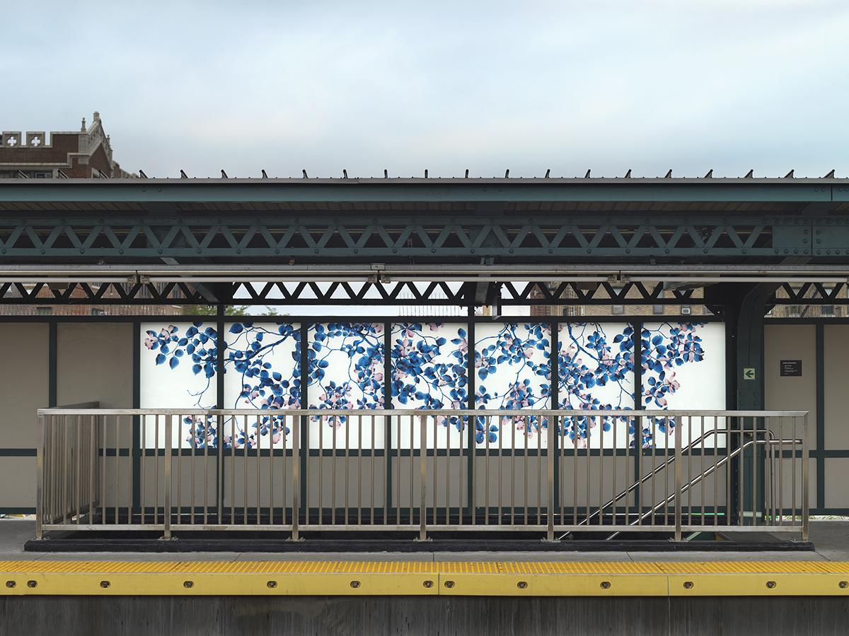 Photograph of the Bay Parkway station platform, with a straight-on view of laminated glass artwork panels installed on the station platform. Panels feature branches with blue and pink leaves on a white background.