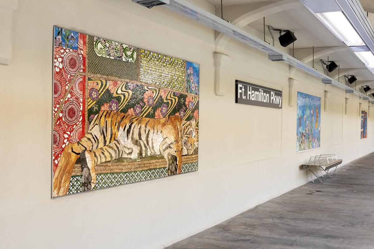 A permanent glass and ceramic mosaic art installation by artist Maria Berrio on the platform walls of the NYCT Fort Hamilton Pkwy station shows two mosaic panels on the platform wall, a resting tiger in the foreground and flowers and butterflies against a blue sky in the background, behind a metal bench. 