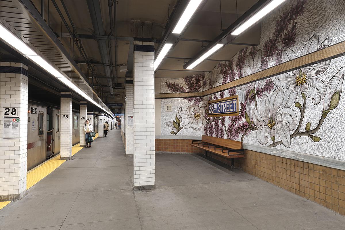 A permanent glass mosaic art installation by artist Nancy Blum on the platform walls of the NYCT 28 St station shows pink and white flowers covering the station right wall with a subway platform in the center and a train on the left.  