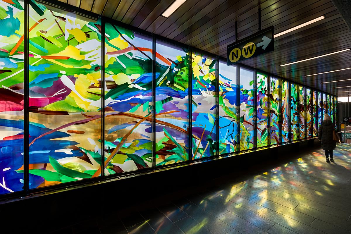 Photo of station mezzanine interior with artwork on the left and a person walking out of station on the right. The artwork is of a nature scene with yellows, greens, browns, blues, and pinks. 