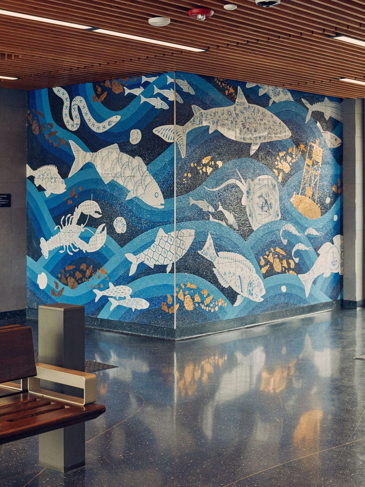 Photograph of an interior of a station with a mosaic on two adjoining walls featuring waves of blue color with sea life swimming throughout. Reflection is captured on the floor and a bench is in the foreground.
