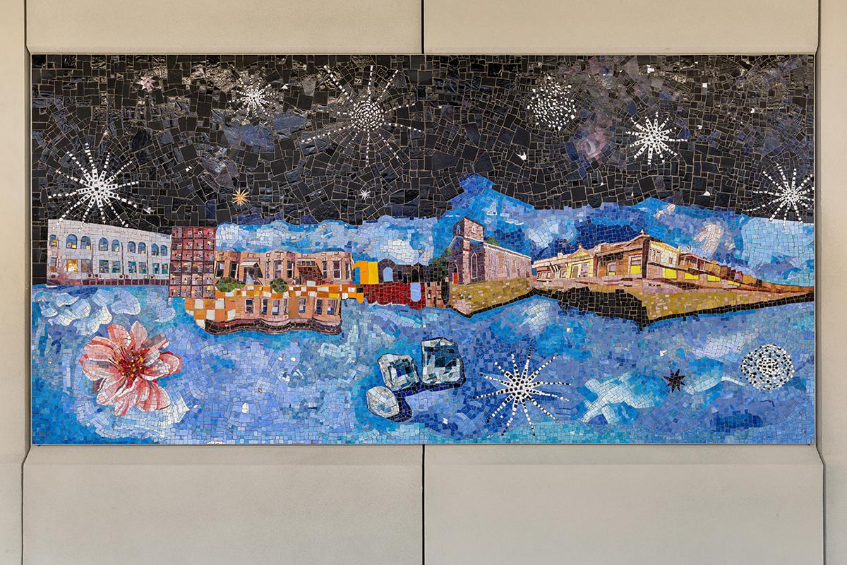 Photograph of a mosaic artwork installed on the platform of the Avenue U station. The mosaic panel has a blue sky and a black ground with objects such as buildings, a pink flower, stars, and ice cubes floating in space. 