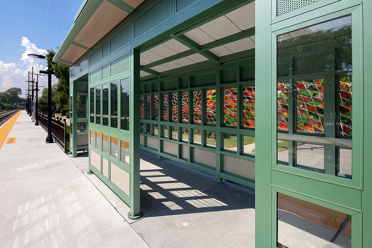 A view of waiting shelter interior as seen from the platform. The shelter features glass artwork with red, green, orange colors framed in geometric forms.