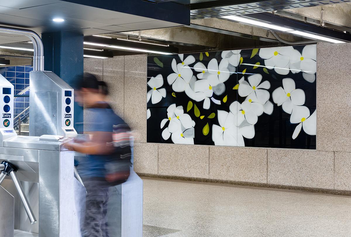 Photograph of laminated glass artwork on wall of 57th Street station. Large hand painted black and white flowers hang on the stone wall of the station, while a blurred person swipes through the turnstile in the foreground.