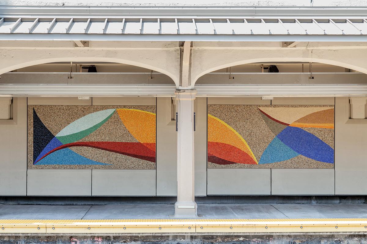 A photograph of glass mosaic artwork on the platform walls of the Bay Parkway station in Brooklyn. Two panels of the colorful abstract artwork with intersecting curved shapes are seen on the platform walls.